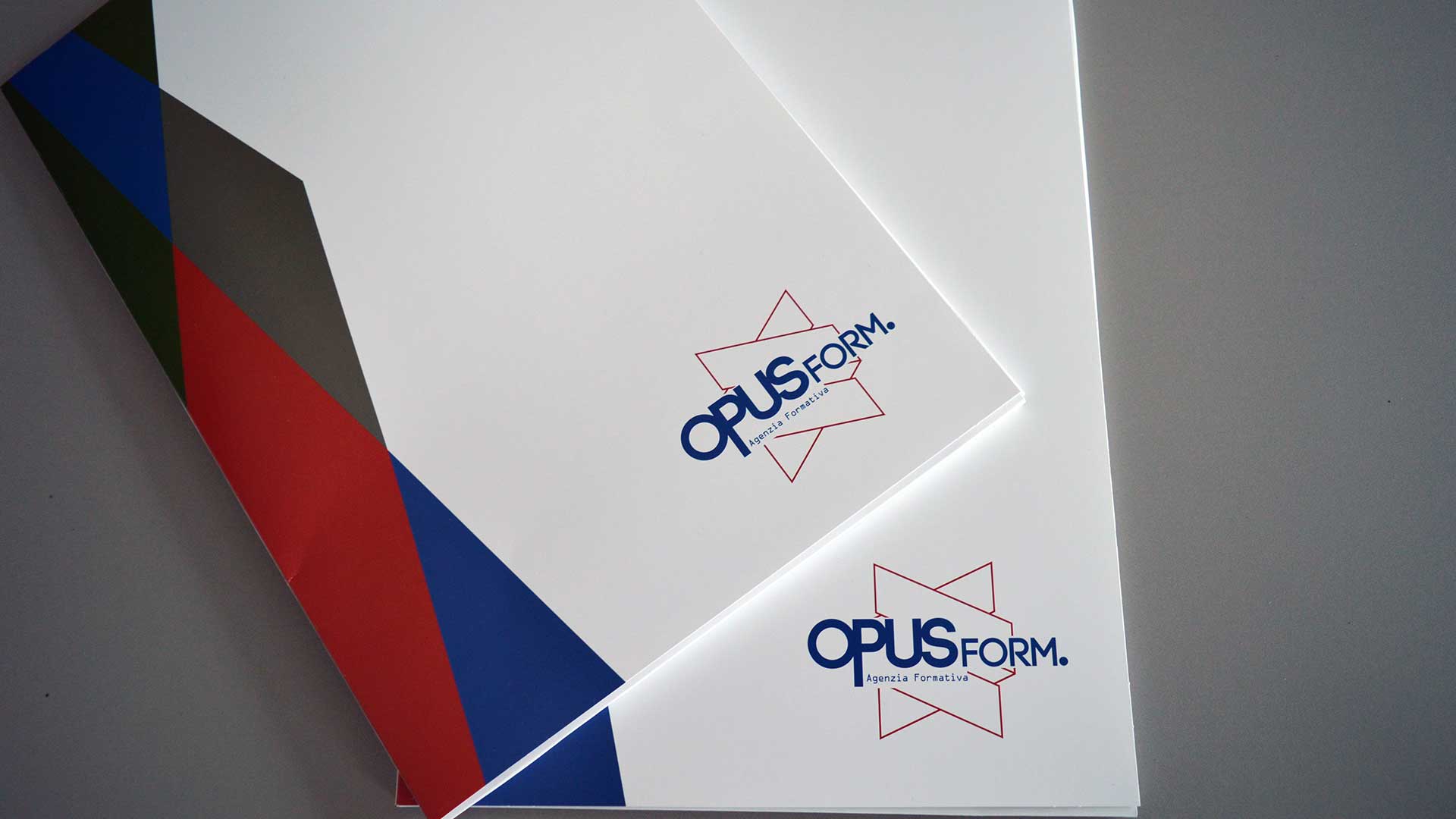 Due cartelline di presentazione bianche con logo OpusForm. Agenzia Formativa e dettagli geometrici colorati sul bordo, sovrapposte su sfondo grigio.