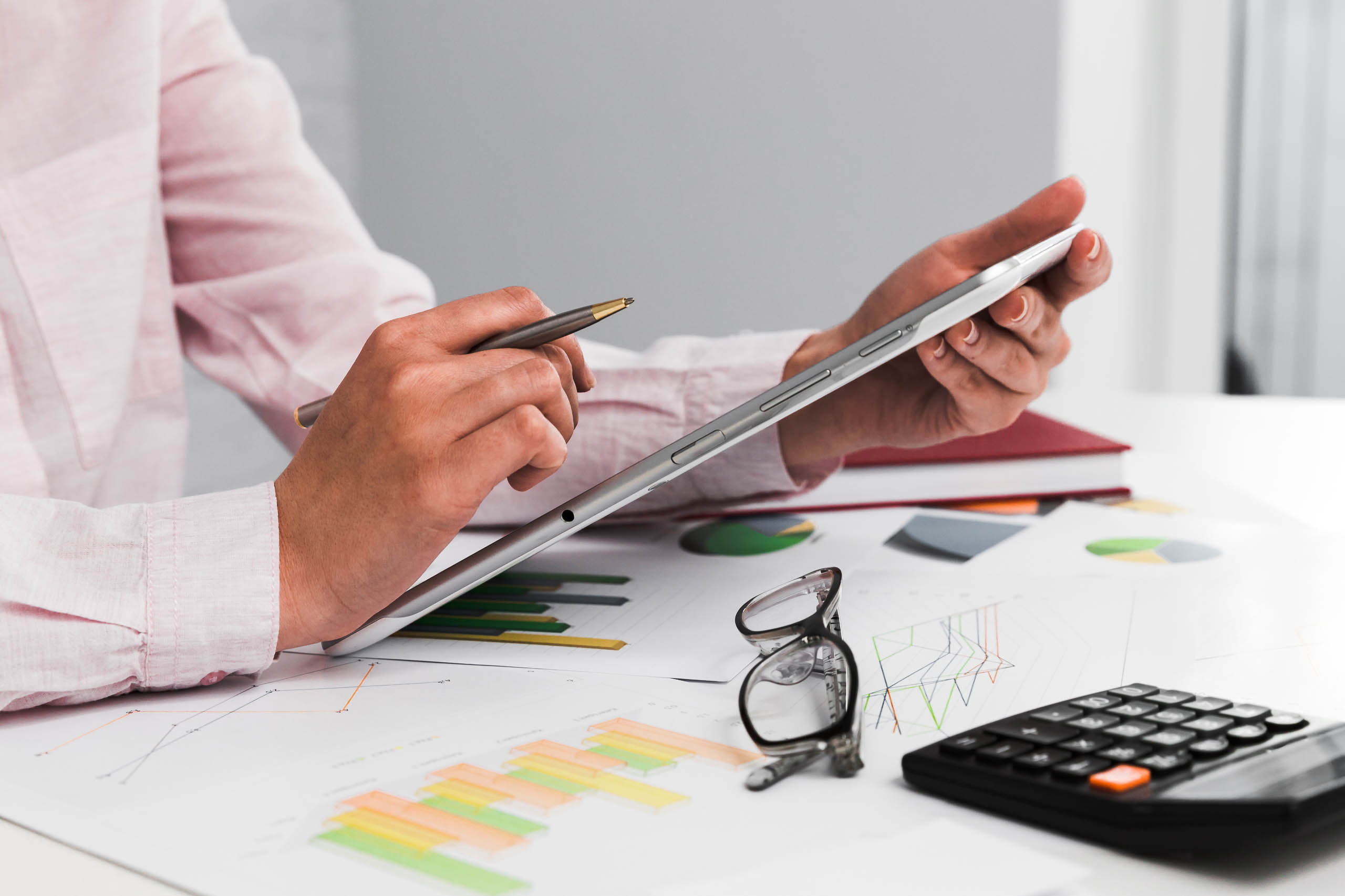Primo piano delle mani di un'analista che utilizza un tablet digitale per consultare grafici e statistiche finanziarie su una scrivania con calcolatrice e occhiali.