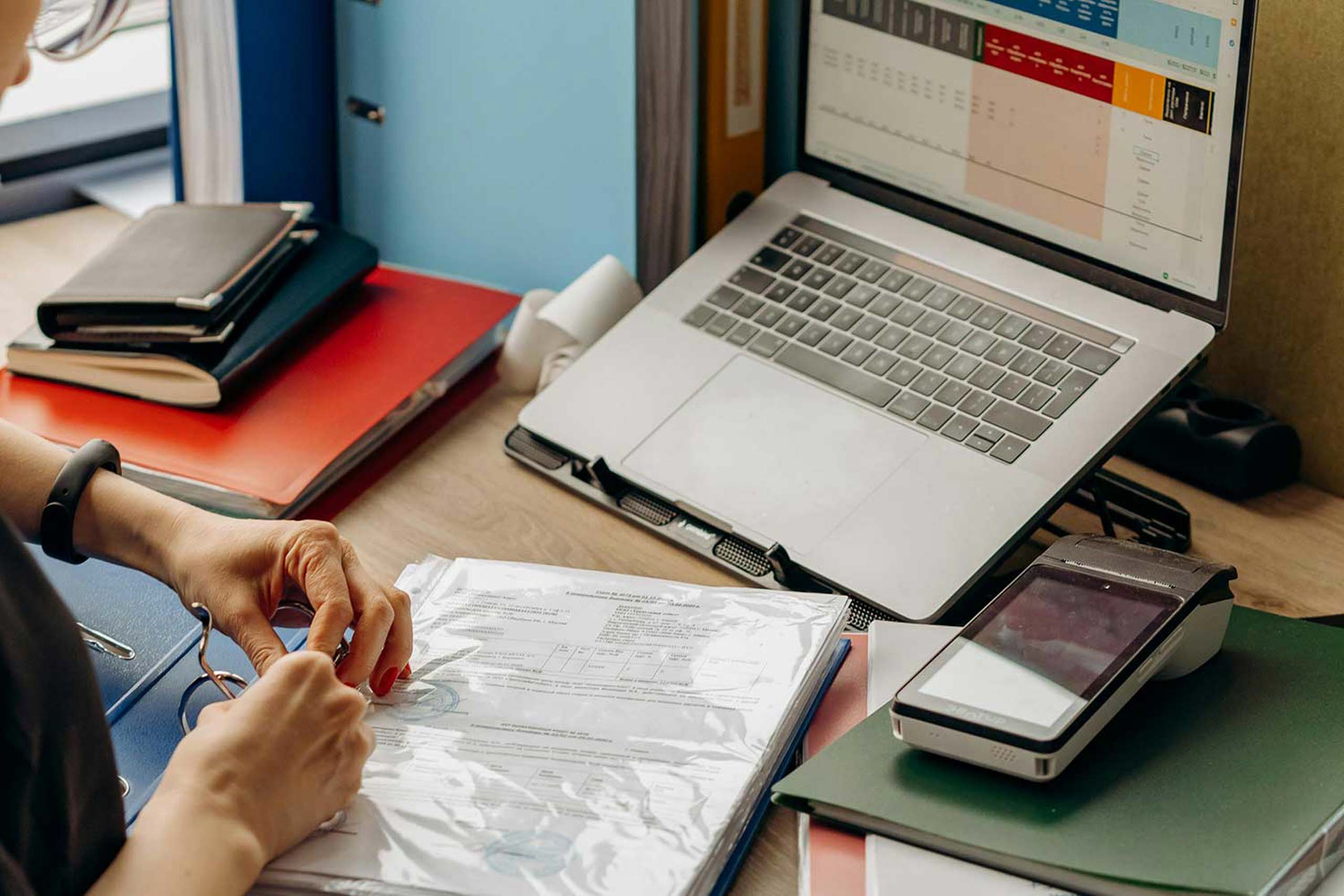 Mani di un'impiegata che consulta documenti contabili in un raccoglitore ad anelli su una scrivania con laptop e terminale POS.