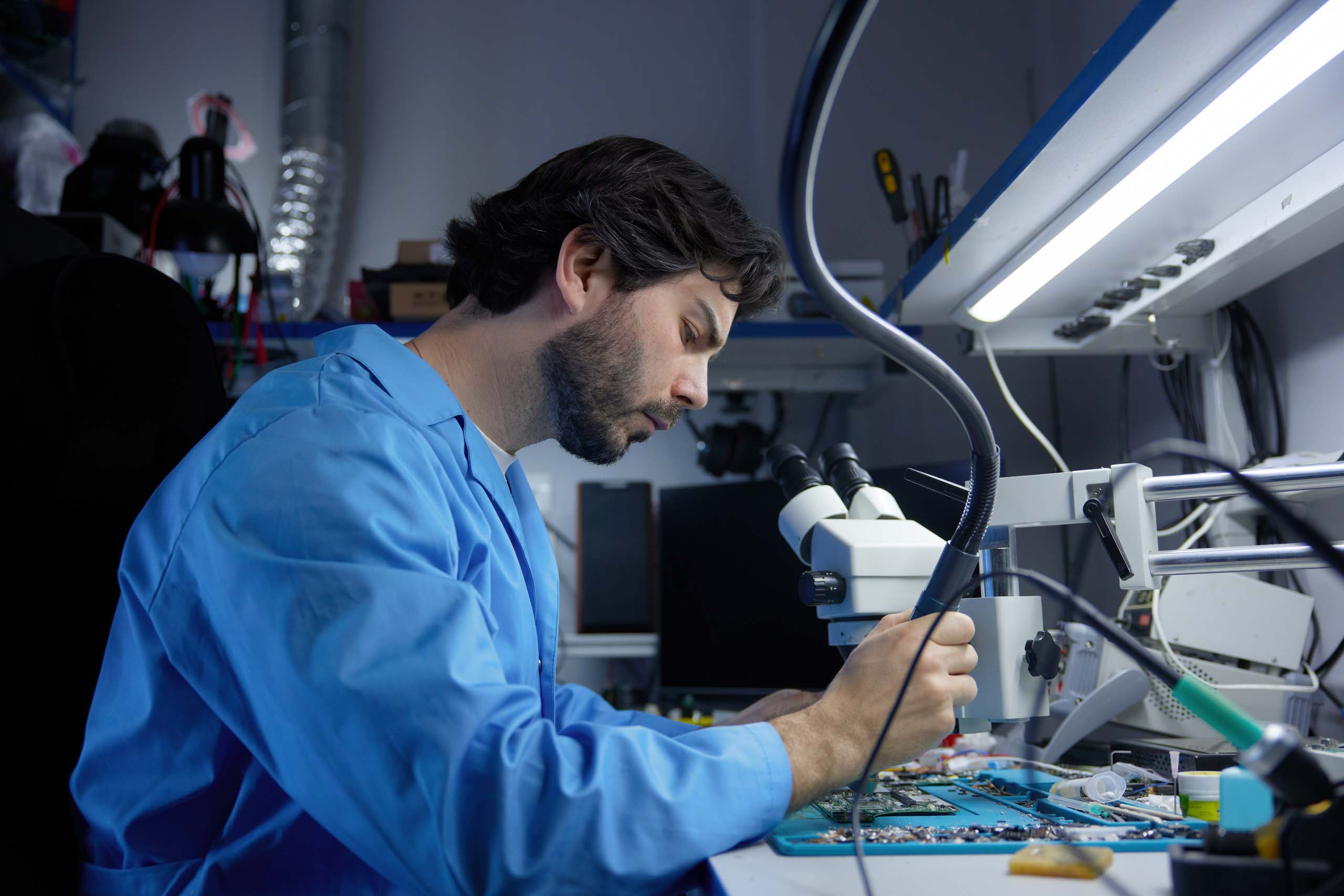 Tecnico meccatronico con camice blu intento a riparare una scheda madre utilizzando un microscopio di precisione e un saldatore in laboratorio.