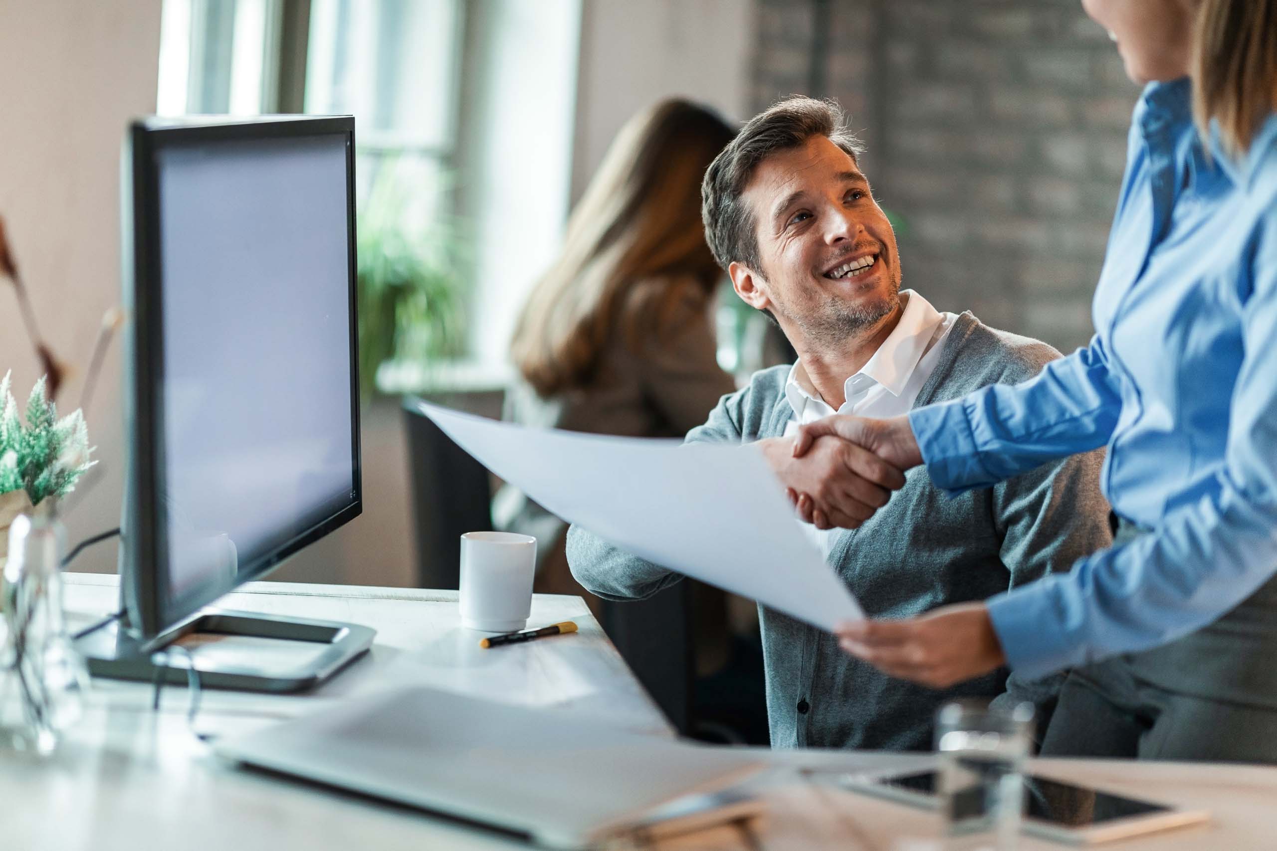Un professionista sorridente stringe la mano a una collega alla scrivania al termine di un colloquio con un documento in mano, simbolo di accordo e di inserimento lavorativo di successo con OpusForm.