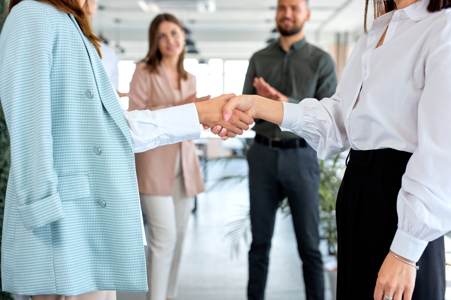 Stretta di mano tra due donne d'affari in ufficio con colleghi che applaudono sullo sfondo per celebrare un accordo.