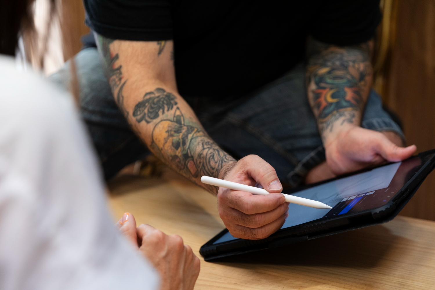 Tatuatore con braccia tatuate che illustra un progetto di tatuaggio su un tablet digitale a un cliente durante una seduta di consulenza.